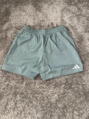 Ljusgråa träningsshorts från Adidas - Snygga ljusgråa träningsshorts från Adidas med elastisk midja och diskret logga på vänster ben. Shortsen har en enkel och sportig design, perfekta för gymmet eller löprundan. Tillverkade i lätt och snabbtorkande syntetmaterial.