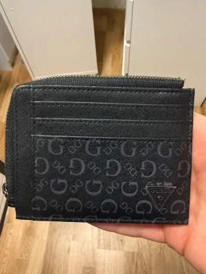 Svart plånbok från Guess med dragkedja - Snygg svart plånbok från Guess med dragkedja runt kanten. Ena sidan har flera kortfack och Guess-logga i metall, andra sidan är i blankt material med präglad logga. Plånboken har ett mönster med G-bokstäver och är tillverkad i konstläder.
