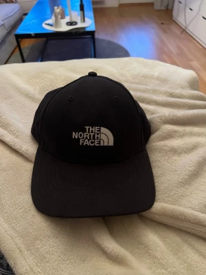 Svart keps från The North Face - Säljer en svart keps från The North Face med klassisk böjd skärm och justerbar rem bak. Kepsen har broderad vit logga framtill och är tillverkad i ett slitstarkt tyg. Perfekt för dig som vill ha en stilren och enkel accessoar.
