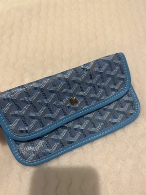 Blå plånbok med mönster - Plånboken har blå kant, tryckknapp i metall och insida i ljust tyg. Perfekt storlek för kort och sedlar, och riktigt trendigt mönster.