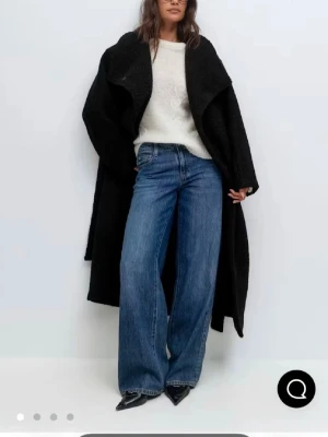 Lurvig jacka ifrån Gina med prislapp - Stilren svart oversized kappa med bred, markerad krage och långa ärmar. Kappan har en avslappnad passform och faller snyggt ned till anklarna. Perfekt för dig som gillar en trendig och minimalistisk look.