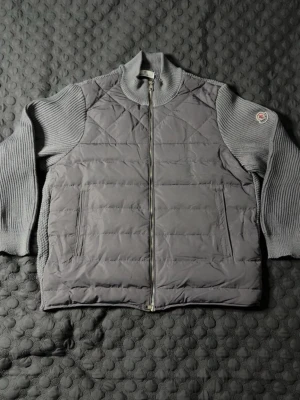 SJUKT BILLIG stickad cardiganjacka Moncler  - Snygg grå cardiganjacka från Moncler med quiltad front och stickade ärmar. Jackan har hög krage, dragkedja framtill och Moncler-logga på ärmen. Perfekt för lager-på-lager och en clean look. STORLEK M, PASSAR M. HELT NY, INGET TECKEN PÅ SLITAGE.