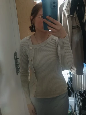 Beige ribbad blus med knappar - Trendig beige ribbad blus med bred fyrkantig ringning och dekorativa stora knappar längs sidan. Figurnära modell med långa ärmar som ger en stilren och modern look. Perfekt att matcha med både kjol och byxor för en snygg outfit.