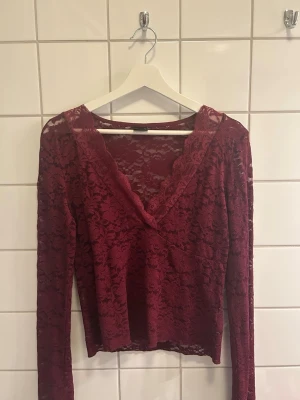 Vinröd spetsblus med v-ringning - Vinröd långärmad blus i genombruten spets med v-ringad hals. Blusen har ett feminint och elegant uttryck med blommigt spetsmönster och är lätt transparent. Perfekt för dig som vill ha en trendig och snygg topp med det lilla extra.