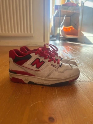 New Balance 550 vita/röda sneakers - Säljer ett par New Balance 550 sneakers i vitt och rött med röda skosnören och detaljer. Skorna har en låg siluett, perforerat läder på ovandelen och tydlig New Balance-logga på sidan. Snygg retrostil som passar till många outfits. små defekter i skinnet syns i bild 
