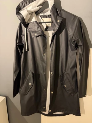 Svart regnjacka med huva från 157 - Snygg svart regnjacka från 157 med vit insida och dragsko i huvan. Jackan har tryckknappar framtill, två fickor med lock och långa ärmar. Perfekt för regniga dagar och enkel att matcha med olika outfits.