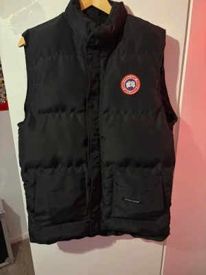 Canada goose väst  - Snygg svart dunväst från Canada Goose med klassisk logga på bröstet. Västen har hög krage, två stora framfickor och stängs med knappar och dragkedja. Perfekt för lager på lager och håller dig varm under kyliga dagar.