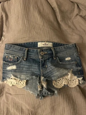 Blå jeansshorts med spets från Hollister low waist - Säljer ett par blå jeansshorts från Hollister med slitna detaljer och råa kanter. Shortsen har vit spets längs bensluten och klassisk femficksdesign. Perfekta för sommaren och ger en avslappnad, trendig look.