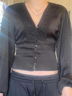 Svart blus med spetsdetaljer - Stilren svart blus med v-ringning och dekorativa spetsdetaljer längs urringningen. Blusen har knappar framtill och är tillverkad i ett glansigt, mjukt material som ger en elegant look. Långa ärmar med lätt puff och smal passform i midjan.
