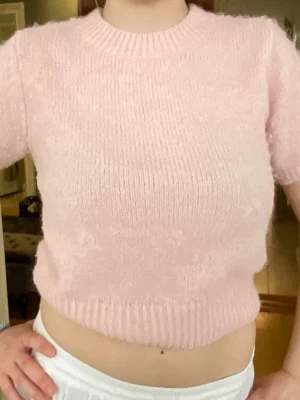 Ljusrosa stickad kortärmad tröja - Supersöt ljusrosa stickad tröja med korta ärmar och rund hals. Tröjan har en mjuk, fluffig känsla och ribbstickade kanter vid hals, ärmslut och nederkant. Perfekt för dig som gillar en feminin och trendig look.