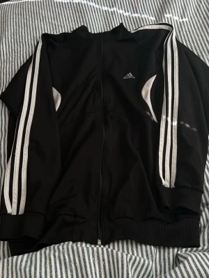 Svart Adidas träningsjacka - Säljer en svart träningsjacka från Adidas med klassiska vita ränder längs ärmarna och dragkedja framtill. Jackan har ribbade muddar och vita detaljer vid sidorna. Perfekt för sport eller chill. Materialet är troligtvis polyester.