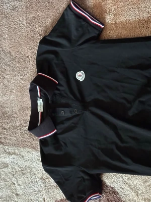 Svart Moncler pikétröja med ränder - Klassisk svart pikétröja från Moncler med vit och röd rand på krage och ärmslut. Tröjan har knappslå framtill och Moncler-logga broderad på bröstet. Perfekt till jeans eller shorts för en clean look.