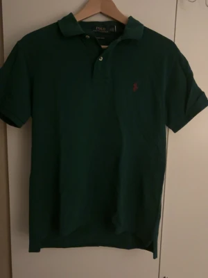 Grön piké från Polo Ralph Lauren - Klassisk grön pikétröja från Polo Ralph Lauren med röd broderad logga på bröstet. Modellen är custom slim fit och har korta ärmar samt krage med knappar. Perfekt för en stilren och avslappnad look. Topp skick. Priset kan diskuteras. Hör av er vid frågor 
