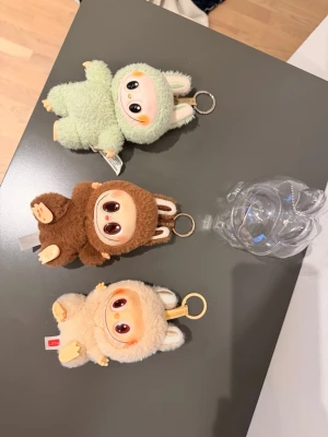 Labubus, Söta nyckelringar med kaninmotiv - Tre fluffiga nyckelringar från POP MART i beige, brun och ljusgrön färg. Varje nyckelring har en gullig figur med stora ögon och kaninöron, tillverkad i mjuk plysch och plast. Perfekt att hänga på väskan eller nyckelknippan för en söt detalj. 3 labubus och 1 transparent fodral. Djur o rökfri hem. Alla i mkt fint skick. 
