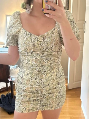 Blommig kortklänning med puffärm - Supersöt kortklänning med småblommigt mönster i ljusa toner. Klänningen har puffärmar, v-ringad urringning och rynkade detaljer längs sidorna. Perfekt för sommarens alla tillfällen och ger en romantisk vibe.