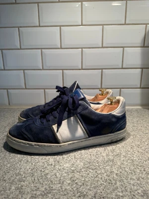Valentino Flycrews Blå - Snygga sneakers från Valentino i marinblå mocka och vitt läder. Skorna har klassisk låg profil, vita och blå detaljer samt matchande blå skosnören. Perfekta för dig som gillar stilrena och tidlösa sneakers. Skorna har sett bättre dagar men är snygga efter en uppfräschning! Ingår dustbags. 