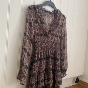 Mönstrad kortklänning från Zara - Supersöt kortklänning från Zara med blommigt och geometriskt mönster i beige, blått och vinrött. Klänningen har långärmade ärmar, v-ringning med volangkant och smockad midja som ger en snygg siluett. Supert skick🩷