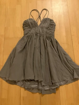 NY  elegant kortklänning med tunna axelband från H&M - Säljer en grå elegant kortklänning med tunna korsade axelband och v-ringad omlott upptill. Klänningen har ett luftigt, svepande tyg och lätt rynkade detaljer över bysten. Perfekt för sommarens eller fest tillfällen. Helt ny med lapparna kvar, storlek 42, H&M