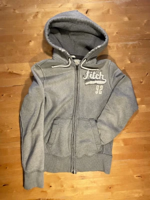 Grå hoodie från Fitch med dragkedja - Snygg grå hoodie från Fitch med broderad logga och siffran 92 på bröstet. Tröjan har dragkedja framtill, två fickor och en stor huva med vita snören. Tillverkad i mjukt sweatshirtmaterial, perfekt för en avslappnad stil.