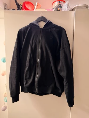 Svart zip hoodie från Gymshark - Säljer en svart hoodie från Gymshark med dragkedja framtill och stor huva. På ryggen finns en stor Gymshark-logga i svart tryck. Hoodien har två fickor fram och ribbade muddar vid ärmslut och nederkant. Perfekt för en sportig och avslappnad stil.