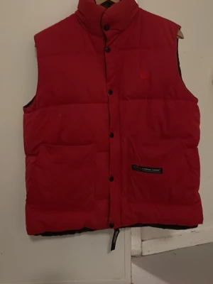 Röd dunväst från Canada Goose - Snygg röd dunväst från Canada Goose med broderad lönnlogga på bröstet och två stora framfickor. Västen har svarta knappar och svart innerfoder. Perfekt för vår och höst. I bra skick, förutom en fläck som är fotograferad. Syns dock inte när man knäpper västen. 
