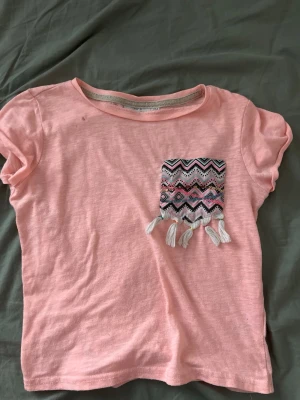 Rosa t-shirt 2016 vibe med mönstrad ficka - Söt rosa t-shirt med korta ärmar och en färgglad mönstrad bröstficka med fransar. Fickan har ett zigzag-mönster i svart, vitt, rosa och turkost samt små paljettdetaljer. Perfekt för en avslappnad och trendig look.