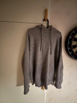 Grå stickad hoodie med huva - Mysig grå stickad hoodie med huva och dragsnören. Tröjan har en avslappnad passform, ribbade muddar vid ärmslut och nederkant samt en enkel, stilren design utan tryck eller logga. Perfekt för dig som gillar en clean och bekväm look.