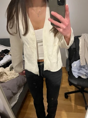 Vit kofta med knappar - Snygg vit kofta med v-ringning och stora knappar framtill. Koftan har två fickor och ribbade muddar vid ärmslut och nederkant. 