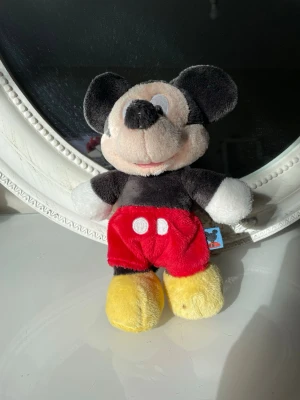 Mjukisdjur Mickey Mouse Disney - Söt vintage mjukisdjur av Mickey Mouse från Disney. Gosedjuret har svart kropp, röda byxor med vita knappar, gula skor och stora runda öron. Tillverkad i mjukt plyschmaterial och har en liten svans baktill. Perfekt som present eller samlarobjekt.