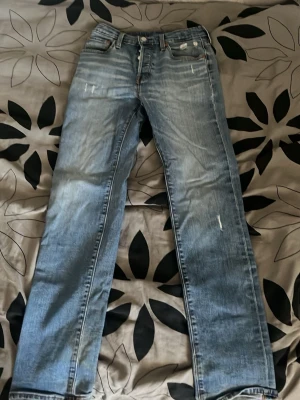 Ljusblå raka jeans från Levi's - Säljer ett par ljusblå jeans från Levi's med raka ben och klassisk femficksmodell. Jeansen har slitningar på låren och knäna för en trendig, avslappnad look. Tillverkade i denim med Levi's ikoniska läderpatch bak i midjan.