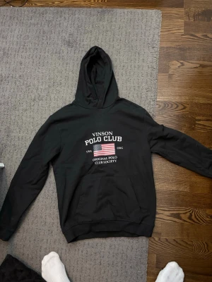 Svart hoodie från Vinson Polo Club - Snygg svart hoodie från Vinson Polo Club med tryck framtill i vitt och amerikansk flagga. Klassisk känguruficka och huva. Perfekt för en avslappnad och sportig stil. Mjuk bomullskänsla och ribbade muddar vid ärmar och nederkant.
