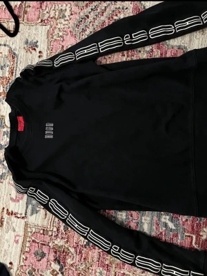 Svart sweatshirt från Hugo - Svart sweatshirt från Hugo med vit logga på bröstet och coolt vitt tryck längs båda ärmarna. Tröjan har rund halsringning och är tillverkad i mjuk bomull. Perfekt för en avslappnad och trendig stil.