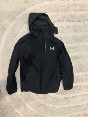 Svart vindjacka från Under Armour - Snygg svart vindjacka från Under Armour med huva och dragkedja framtill. Size M priset kan diskuteras.