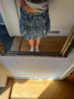 Blå paisleymönstrad kortkjol - Supersnygg kortkjol med paisleyinspirerat mönster i blått, brunt och vitt. Kjolen har resår i midjan och volangdetalj som ger en avslappnad och trendig look. 