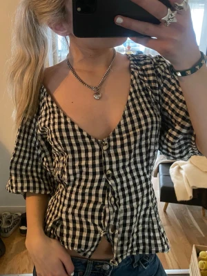 Rutig blus med puffärm och knappar - Snygg svartvit rutig blus med puffiga korta ärmar och djup v-ringning. Blusen har knäppning framtill och lätt figursydd passform som ger en feminin siluett. Perfekt till jeans eller kjol för en trendig look.