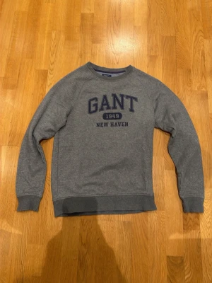 Grå sweatshirt från GANT - Klassisk grå sweatshirt från GANT med tryckt logga och texten 'GANT 1949 NEW HAVEN' på bröstet. Tröjan har rund halsringning, ribbade muddar vid ärmslut och nederkant samt en avslappnad passform. Perfekt för en casual och sportig stil.