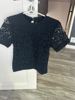 Svart spetsblus med kort ärm från Lindex  - Snygg svart blus i spets med korta ärmar och rund hals. Blusen har ett detaljerat bladmönster i spetsmaterial som ger en elegant look. Perfekt att styla med jeans eller kjol för en trendig outfit.