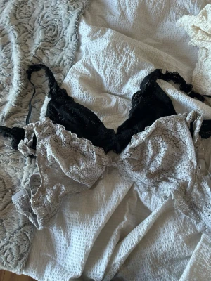 Två spetsbhar i svart och beige - Säljer två bhar i spets, en i svart och en i beige. Båda har tunna axelband och dekorativa volangkanter. Perfekta för dig som gillar romantisk och feminin stil. Inget vadderat och utan bygel för extra komfort.