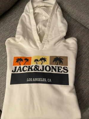 Vit hoodie från Jack & Jones.Barn strl 176 - Snygg vit hoodie från Jack & Jones med tryck av palmer och texten 'Los Angeles, CA' på bröstet. Färgglada detaljer i orange, gul och brun ger en cool retrovibe. Mjuk och bekväm med huva, perfekt för chill dagar.strl 176