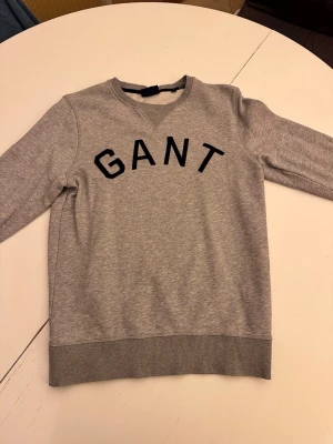 Grå sweatshirt från GANT - Snygg grå sweatshirt från GANT med svart broderad logga över bröstet. Tröjan har rund halsringning, ribbade muddar vid ärmslut och nederkant samt en klassisk relaxed fit. Perfekt till jeans eller joggers.