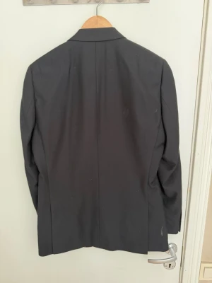 Svart klassisk kavaj med knappar - Stilren svart kavaj med två knappar framtill och klassiska slag. Kavajen har tre fickor på framsidan och fyra dekorativa knappar vid ärmsluten. Perfekt för en dressad look och enkel att matcha med skjorta eller t-shirt.
