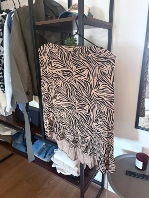 Beige zebramönstrad långkjol - Säljer en beige långkjol med svart zebramönster och volangkant nertill. Kjolen har en flowy passform och är perfekt för dig som vill sticka ut med ett djurmönster. Supersnygg att matcha med en enkel topp eller stickad tröja.