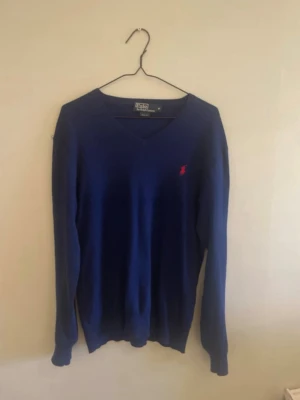 Blå stickad tröja Polo Ralph Lauren - Mörkblå stickad tröja från Polo Ralph Lauren i slim fit-modell. Tröjan har V-ringning, långa ärmar och den klassiska röda logotypen broderad på bröstet. Perfekt för dig som gillar stilrena och tidlösa plagg.