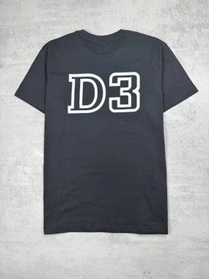 Svart Nikon D3 t-shirt - Svart t-shirt från Harvest med stort vitt D3-tryck på framsidan och en liten gul Nikon-logga på bröstet. Klassisk passform med rund hals och korta ärmar. Perfekt för dig som gillar foto och vill ha en stilren look.