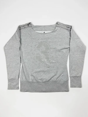 Grå stickad tröja från Adidas - Snygg grå stickad tröja från Adidas med diskret mönster av små trianglar framtill. Tröjan har rund halsringning, långa ärmar och dragkedjedetaljer på axlarna för en modern touch. Perfekt för en avslappnad och sportig stil.