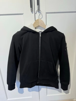 Svart hoodiejacka från Moose Knuckles - Säljer en svart hoodie från Moose Knuckles med dragkedja och två fickor framtill. Insidan är fodrad med vit, mjuk fleece och ärmen har den ikoniska logotypen i metall. Perfekt för dig som vill ha en stilren och bekväm hoodie med coola detaljer