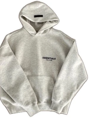 Essentials Fear of God hoodie - Snygg grå hoodie från Essentials Fear of God med svart logga på bröstet och stor text på ryggen. Klassisk oversized passform, stor huva och magficka. Tillverkad i mjukt bomullsmaterial som känns riktigt skönt mot huden. Perfekt för en avslappnad streetwear-look.