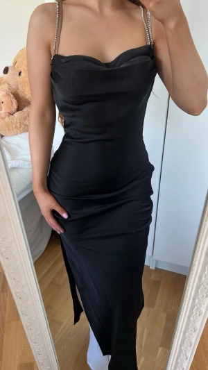 Klänning - Säljer en elegant svart långklänning med smala, glittriga axelband och draperad urringning. Klänningen har en figurnära passform och en slits på sidan för extra stilpoäng. Perfekt för dig som vill sticka ut på nästa event! Storlek XS/S, ny med prislapp kvar då den tyvärr ej kom till användning 🖤