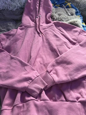 Rosa hoodie med huva - En klassisk rosa hoodie med huva och dragsnören. Hoodien har ribbade muddar vid ärmslut och midja, samt en avslappnad passform. Perfekt för en chill och bekväm stil. Materialet känns mjukt och skönt mot huden.