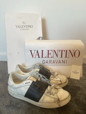 Valentino Skor - Säljer ett par vita sneakers från Valentino Garavani med en bred svart läderdetalj över mitten och klassisk snörning. Skorna har en låg silhuett och är tillverkade i läder med gummisula. Snygg och stilren design som sticker ut.